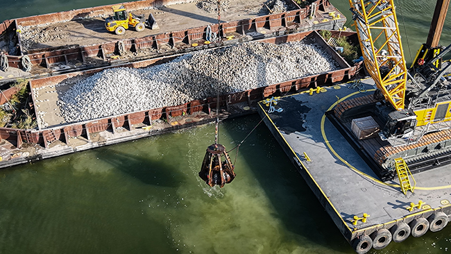 An empty grapple suspended from a crane hovers above water. Nearby is a barge loaded with cobble. The water beneath the crane looks cloudy from cobble that was just deposited in the water.